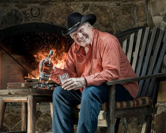 Terry Bradshaw in a rocking chair pouring a glass of Bradshaw Bourbon in front of a stone fireplace.